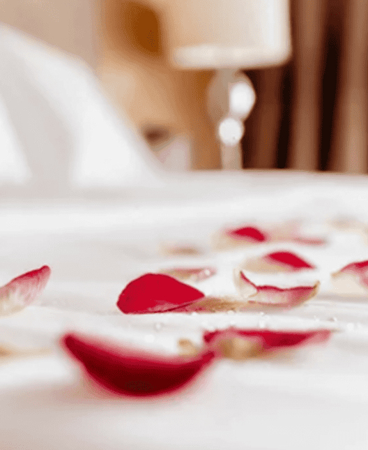 Hotel worth first date Red rose petals scattered on a neatly made white bed in a softly lit hotel room, with a blurred bedside lamp in the background.